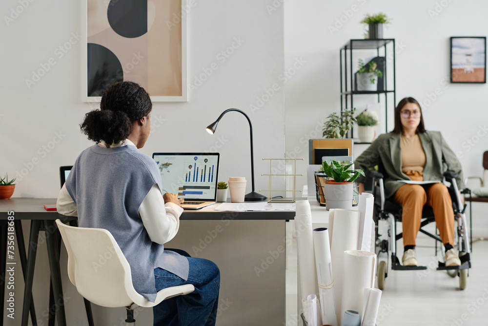 Selective focus shot of unrecognizable Black woman working on laptop in modern office, her colleague with disability using wheelchair in background Selective focus shot of unrecognizable Black woman working on laptop in modern office, her colleague with disability using wheelchair in background