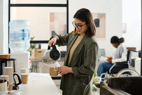 Young woman wearing smart casual outfit with eyeglasses making drip coffee during break at work in modern office photo