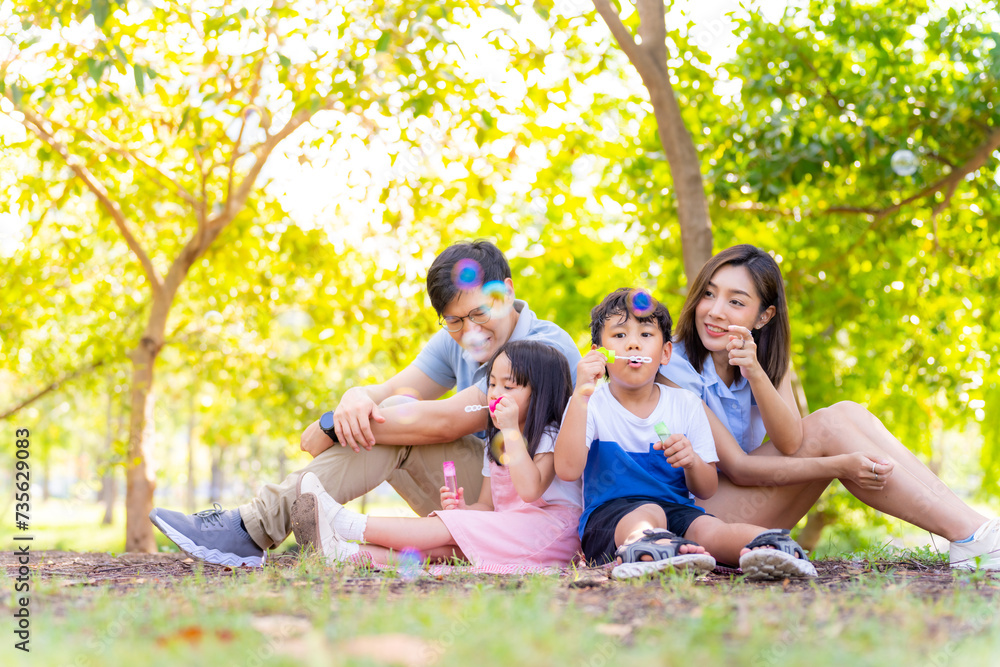 Happy Asian family enjoy and fun outdoor lifestyle on summer holiday vacation. Parents and little child daughter and son spending time playing soap bubble wand together at public park in the city.