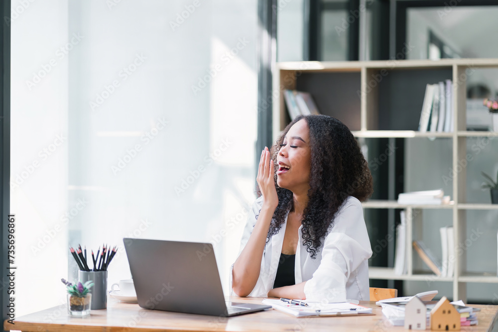 Beautiful Afro American businesswoman using a laptop and yawning while working in a white office. Beautiful Afro American businesswoman using a laptop and yawning while working in a white office.