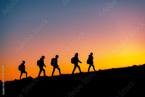 Group of hikers with backpacks silhouettes walks uphill in mountains against sunset sky