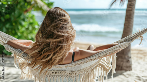 A blonde woman with loose hair relaxes in a hammock on the beach against the backdrop of palm trees and the ocean. © Evgeniia