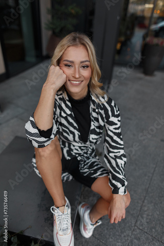 Fashion happy beautiful young girl with a smile in a fashionable zebra print dress and shorts with white sports sneakers sits and relaxes on the street