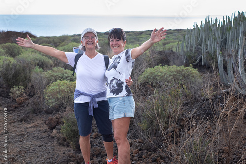 Cheerful couple of senior female friends walking in the nature of Malpais de Guimar at Tenerife island with backpack on shoulder, horizon over sea and sky on background photo