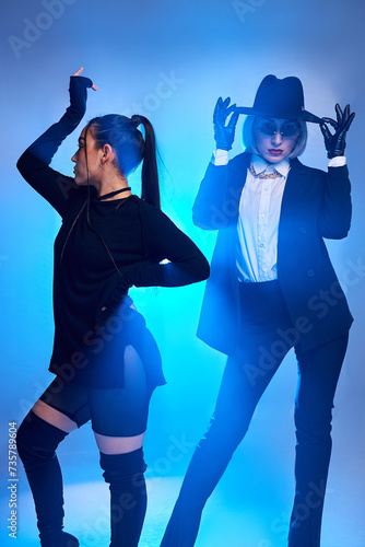 Adorable women in stylish black clothes posing at camera, confident and cool. isolated in studio with blue light in background. european caucasian ladies moving gracefully passionately