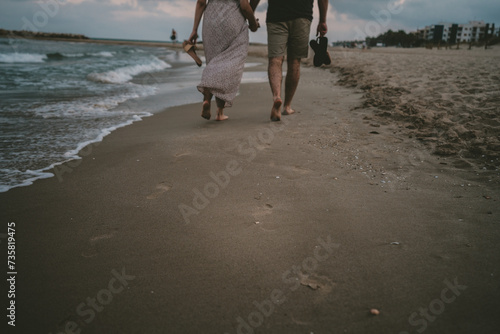 Pareja caminando por la orilla de la playa de espaldas a la cámara. Marcas de huellas en la arena de la orilla con suaves olas del mar al lado. Tienen sus chanclas en las manos. photo