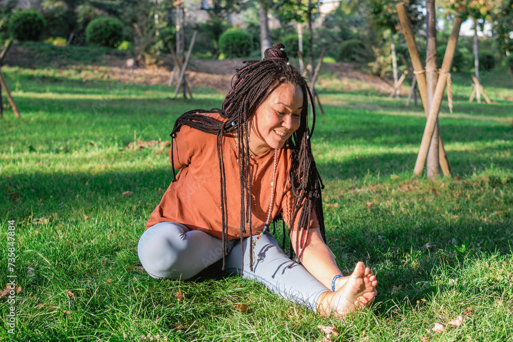 Fototapeta premium Beautiful young woman with long African braids is doing yoga outside in a park. Concept of healthy lifestyle.