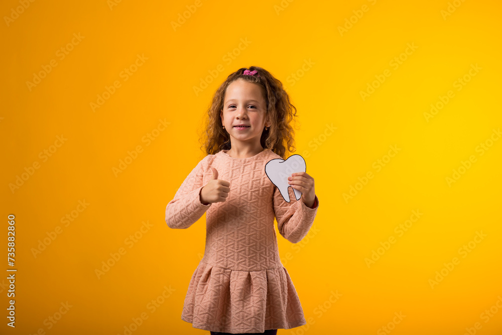 Child girl holding papercraft tooth and showing thumb up gesture over yellow background. Dental health concept Child girl holding papercraft tooth and showing thumb up gesture over yellow background. Dental health concept