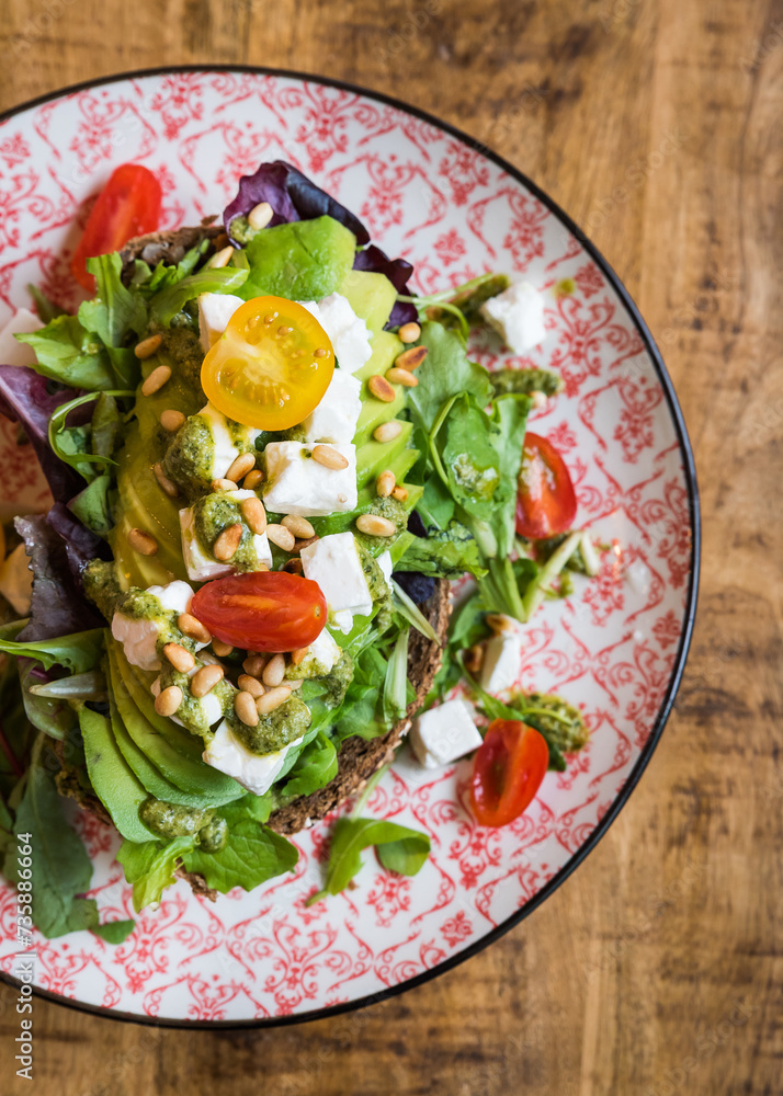 Top view of avocado with feta cheese, tomatoes, pine nuts, pesto and salad on brown bread on plate on wooden table Top view of avocado with feta cheese, tomatoes, pine nuts, pesto and salad on brown bread on plate on wooden table