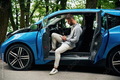 With smartphone in hands. Beautiful young man is sitting in the blue car outdoors © standret