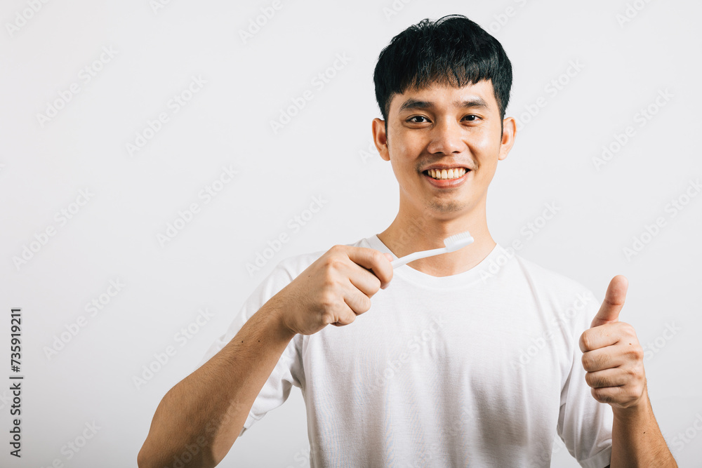Portrait of a smiling Thai young man confidently brushing his teeth and giving a thumbs-up for dental health. Studio shot isolated on white background, promoting dental and oral care. Portrait of a smiling Thai young man confidently brushing his teeth and giving a thumbs-up for dental health. Studio shot isolated on white background, promoting dental and oral care.