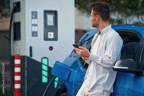 Side view, standing with smartphone, waiting. Man with blue electric car on the charge station