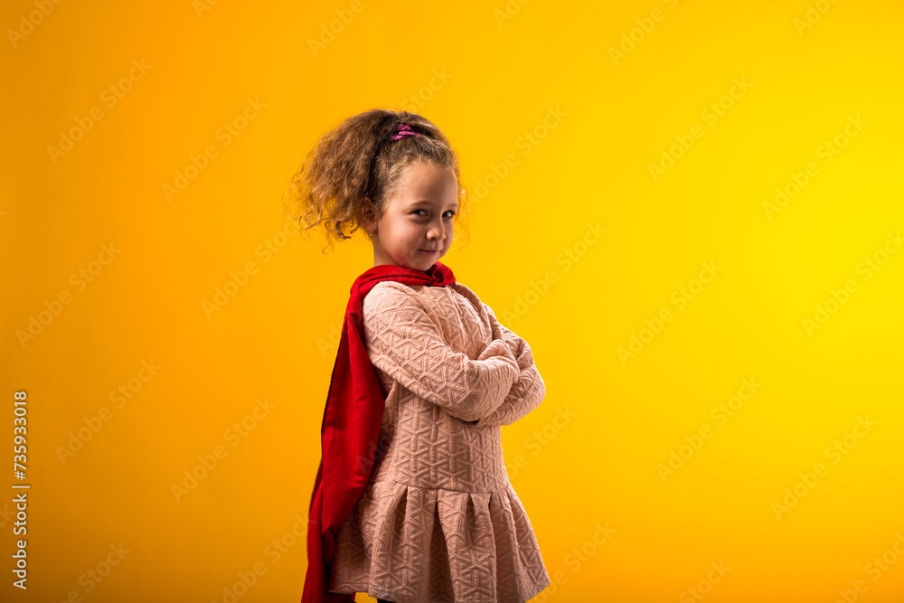 Kid girl superhero in a red cloak on yellow background. Concept of victory and success. Kid girl superhero in a red cloak on yellow background. Concept of victory and success.