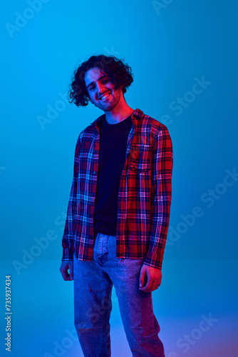 Smiling guy with curly hair, unshaved face, in checkered shirt and jeans smiling against blue background in neon light. Relaxed, casual style for young people. Youth, human emotions, lifestyle concept