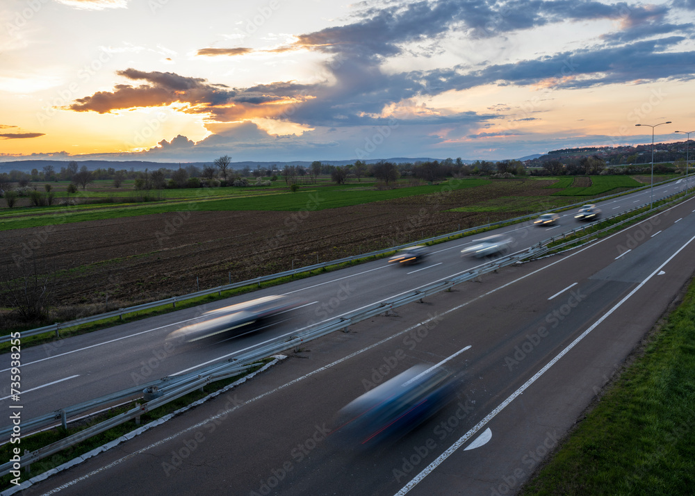 Naklejka premium Cars drive at high speed on the highway through the rural landscape. Fast blurred highway driving. A scene of speeding on the highway. 