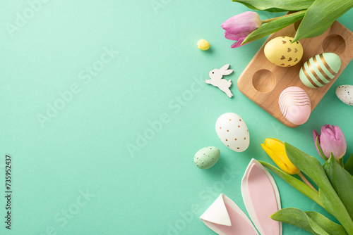 Festive Easter vibes! Top view of color-popping eggs in wooden holder, bunny ears, and tulips. Turquoise backdrop creates a lively atmosphere with space for your Easter wishes or promotional text