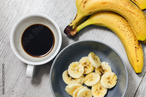 Top view banana salad with coffee. Bowl with fruit slices inside. Banana slices in bowl. Wooden table healthy breakfast. Banana pieces background. White ceramic coffee cup. Morning breakfast.