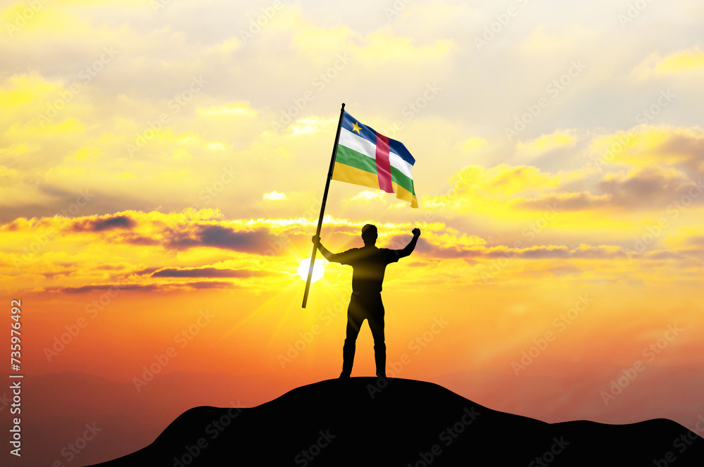 Central African Republic flag being waved by a man celebrating success at the top of a mountain against sunset or sunrise. Central African Republic flag for Independence Day.