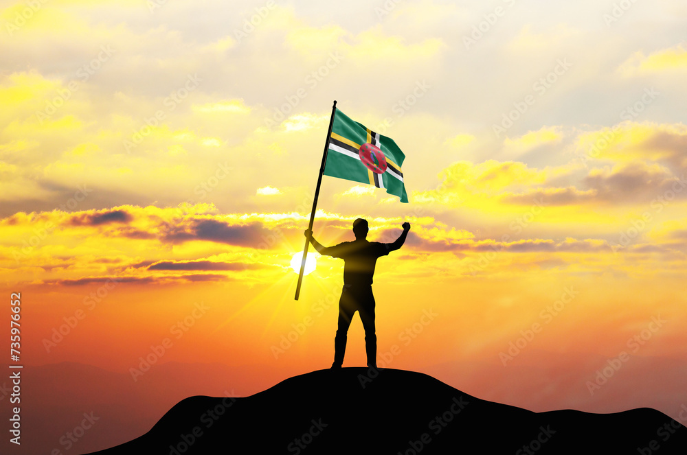 Dominica flag being waved by a man celebrating success at the top of a mountain against sunset or sunrise. Dominica flag for Independence Day.