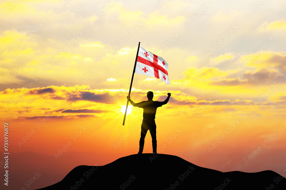 Georgia flag being waved by a man celebrating success at the top of a mountain against sunset or sunrise. Georgia flag for Independence Day.