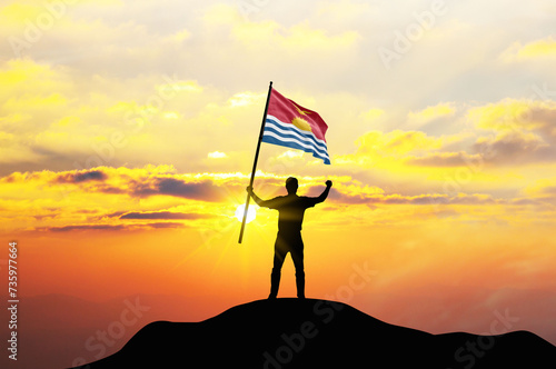 Kiribati flag being waved by a man celebrating success at the top of a mountain against sunset or sunrise. Kiribati flag for Independence Day.