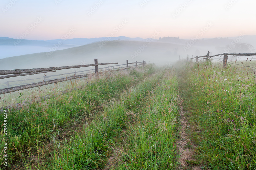 Summer foggy mountain landscape with rural road with fence. Carpathians, Ukraine