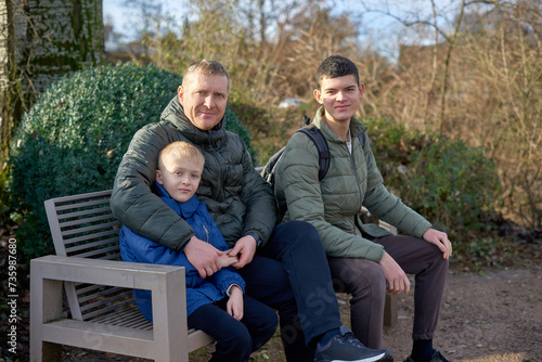 Dad with two sons sitting on a bench in autumn park. Autumnal Family Serenity: Father, 40 Years Old, and Two Sons - Beautiful 8-Year-Old Boy and 17-Year-Old Young Man, Seated in the Park