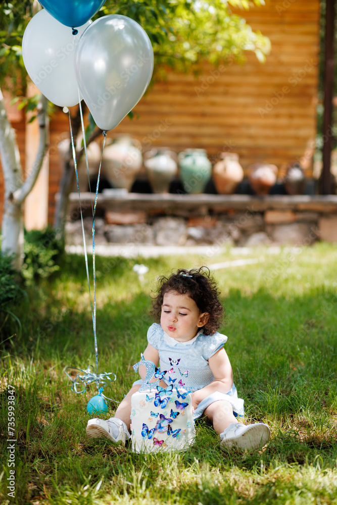 Joyful toddler girl sitting on the grass, next to the balloons and blowing candles on birthday cake