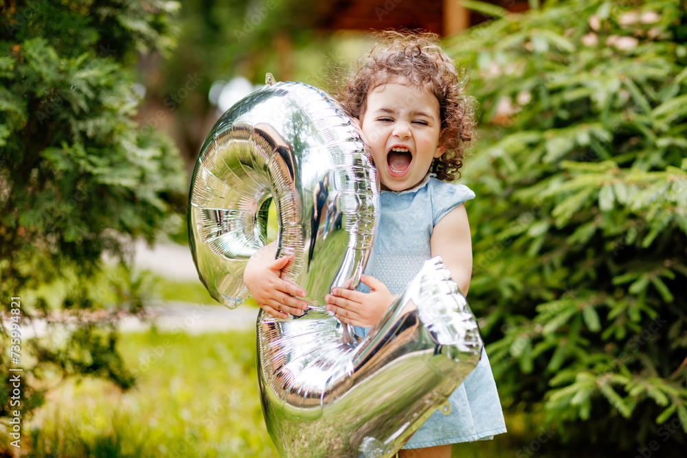 Very happy two years old little girl celebrating her birthday, laughing, holding balloon number