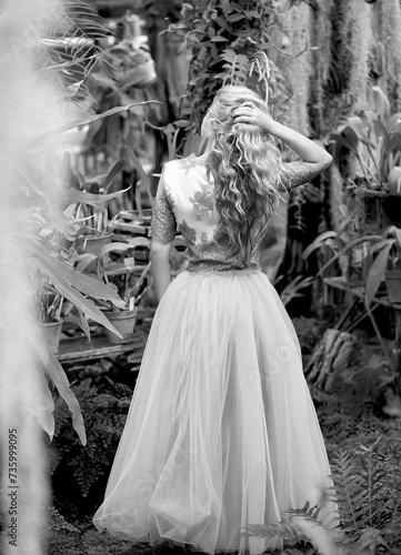 Black and white photo of a stunning girl in a wedding dress, blonde with long curls, view from the back. Summer garden background. Women concept