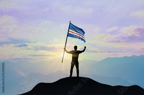 Cabo Verde flag being waved by a man celebrating success at the top of a mountain against sunset or sunrise. Cabo Verde flag for Independence Day.