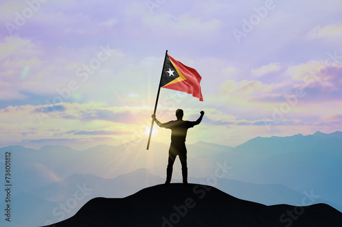 East Timor flag being waved by a man celebrating success at the top of a mountain against sunset or sunrise. East Timor flag for Independence Day.