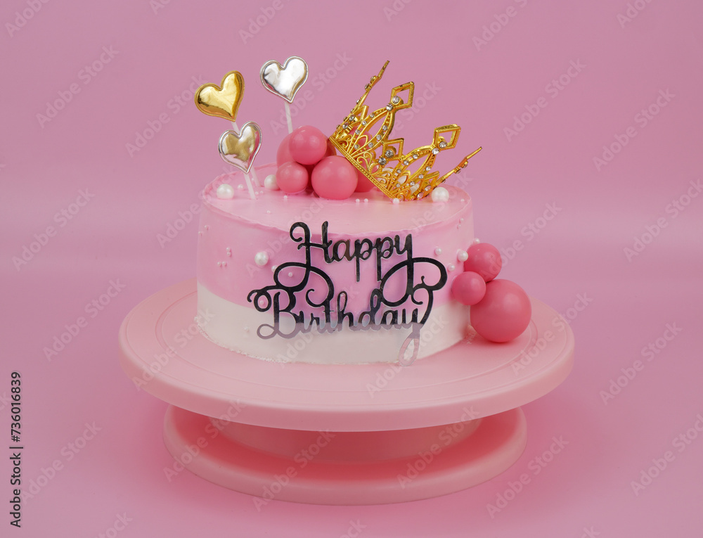 Pink birthday cake with gold crown and hearts on pink round rotating table.	