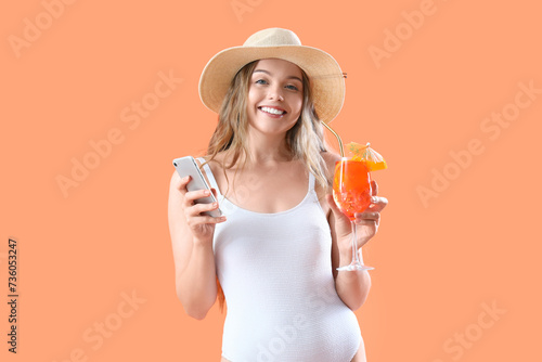Beautiful young woman in swimsuit with glass of tasty aperol spritz and mobile phone on orange background