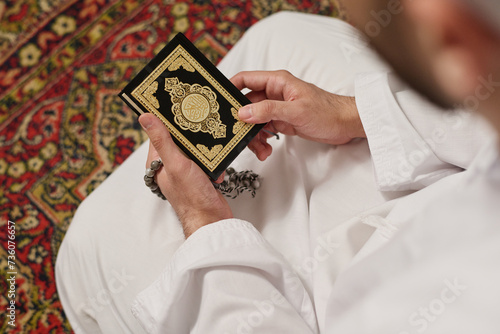 Hands of young Muslim man in white kandora standing on knees on the carpet and holding Holy Quran and rosary beads during silent pray photo