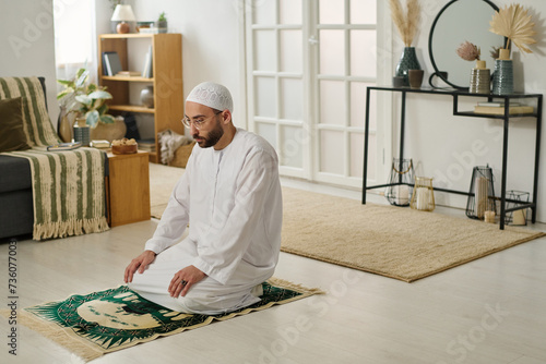 Young religious man in traditional Muslim skullcap and thobe standing on knees on rug in living room and performing silent prayer photo