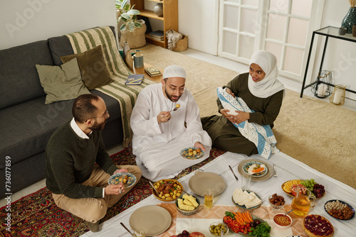 Two young Muslim men sitting on the floor in front of tablecloth with served homemade food, having baked potatoes and talking photo