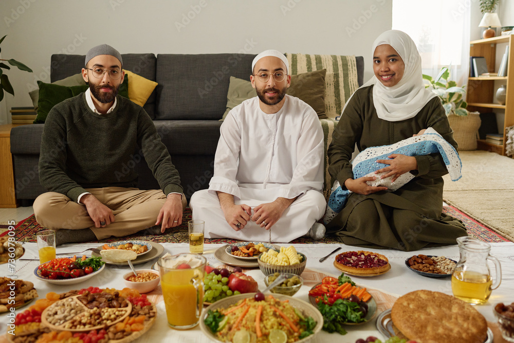 Happy young woman with baby, her husband and their guest looking at camera while sitting on the floor with homemade food on tablecloth Happy young woman with baby, her husband and their guest looking at camera while sitting on the floor with homemade food on tablecloth
