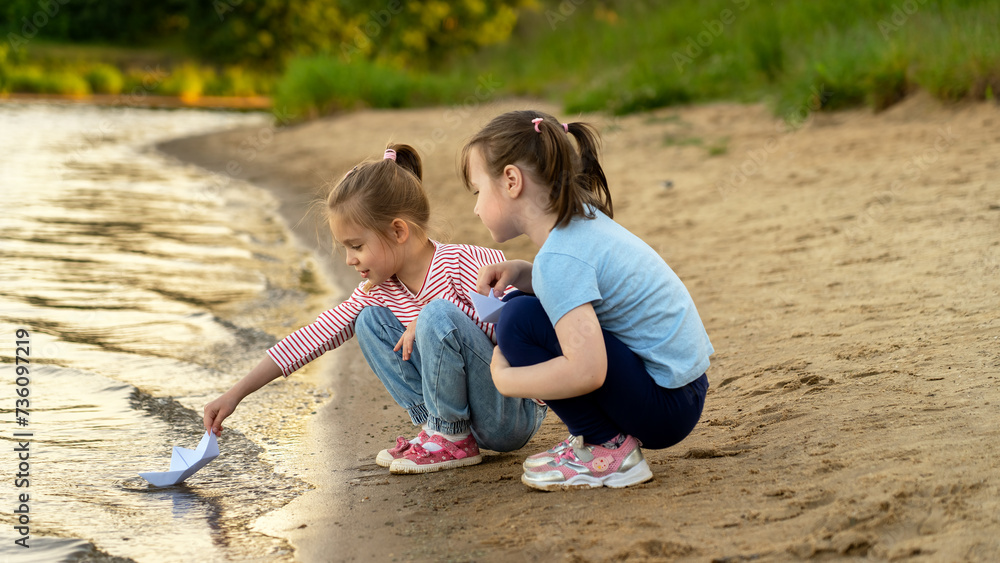 Happy children play with paper boats on river on background of summer park at sunset. Girls walk outdoors by water. Leisure Happy children play with paper boats on river on background of summer park at sunset. Girls walk outdoors by water. Leisure