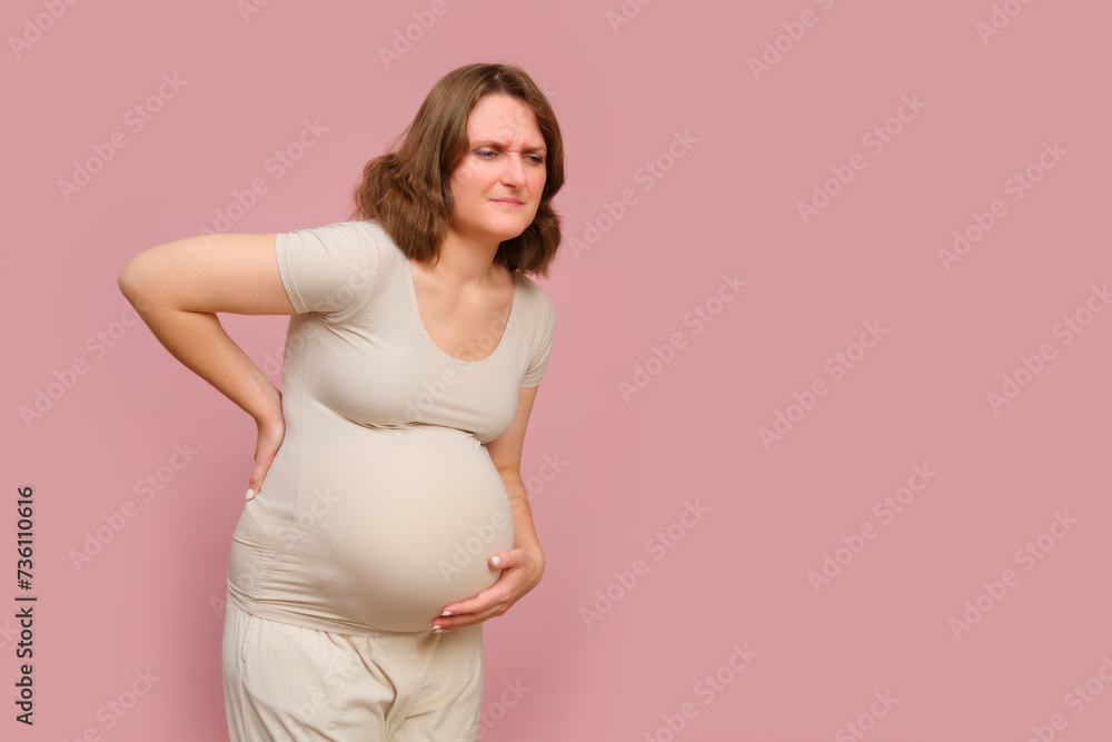 A pregnant woman holds her sore back against a studio pink background. Pregnancy in a woman with a belly, copy space A pregnant woman holds her sore back against a studio pink background. Pregnancy in a woman with a belly, copy space
