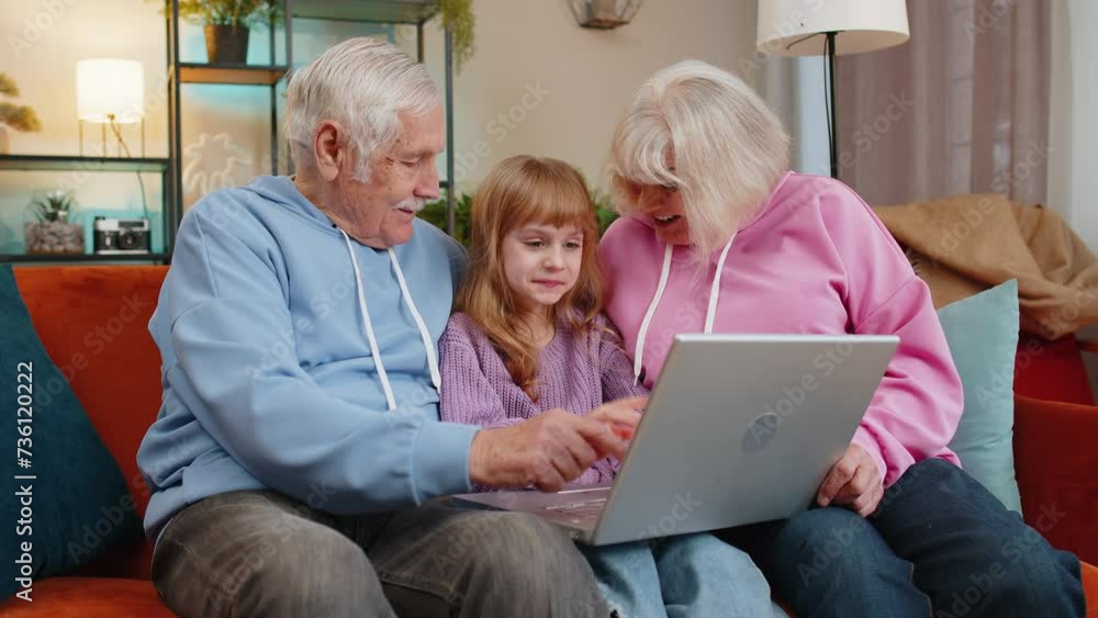Senior Caucasian couple man and woman with child girl granddaughter spending time at home together, sitting on sofa in the living room, using laptop computer. Watching videos playing games on weekend. Senior Caucasian couple man and woman with child girl granddaughter spending time at home together, sitting on sofa in the living room, using laptop computer. Watching videos playing games on weekend.