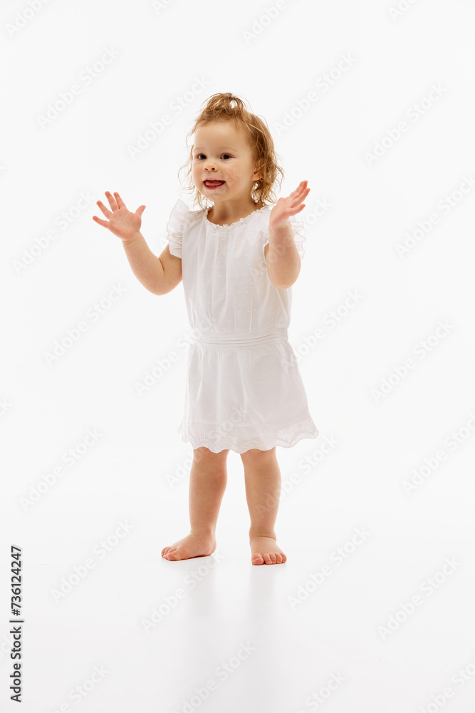 Full-length portrait of cute and happy little baby girl in white dress running and clapping hands of joy against white studio background. Concept of childhood, motherhood, life, birth. Copy space. Ad Full-length portrait of cute and happy little baby girl in white dress running and clapping hands of joy against white studio background. Concept of childhood, motherhood, life, birth. Copy space. Ad