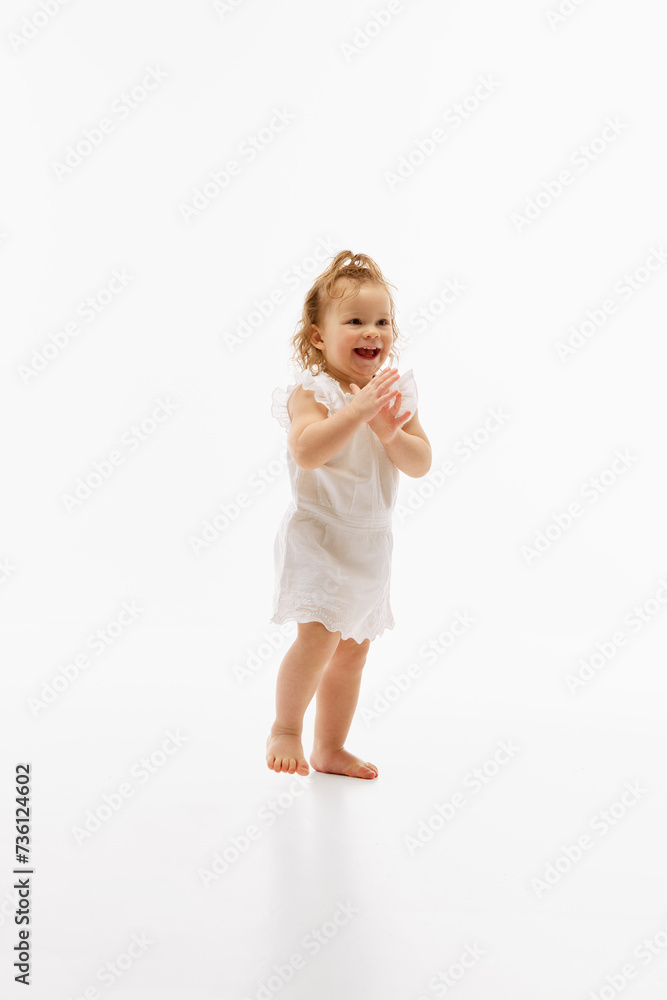 Full-length portrait of smiling little baby girl in white dress running and clapping hands of joy against white studio background. Concept of childhood, motherhood, life, birth. Copy space for ad Full-length portrait of smiling little baby girl in white dress running and clapping hands of joy against white studio background. Concept of childhood, motherhood, life, birth. Copy space for ad