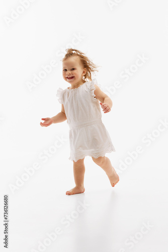 Playful, cute little girl with wet, curly hair running in white dress against white studio background. Baby fashion. Concept of childhood, motherhood, life, birth. Copy space for ad