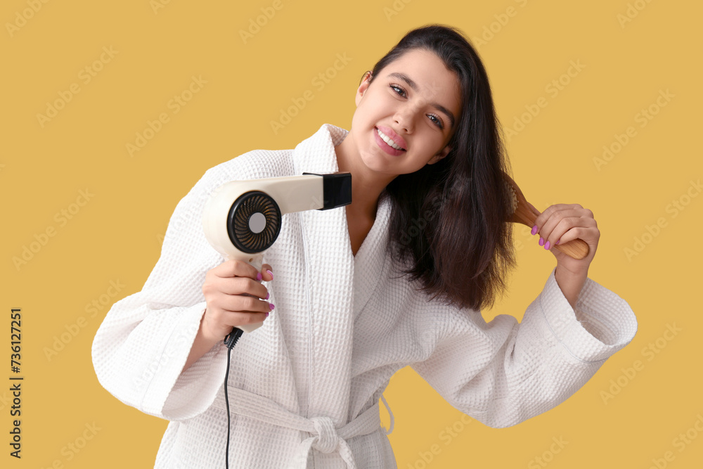 Beautiful young happy woman in bathrobe with hair dryer and brush on yellow background Beautiful young happy woman in bathrobe with hair dryer and brush on yellow background