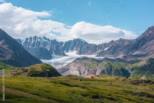 Colorful landscape with orange tent in alpine valley with view to big glacier tongue and large snow-capped mountain range under clouds in blue sky. Green hills and rocks against ice and sheer crags.
