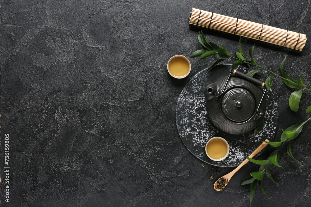 Teapot, cups of hot tea and spoon with dry leaves on black background
