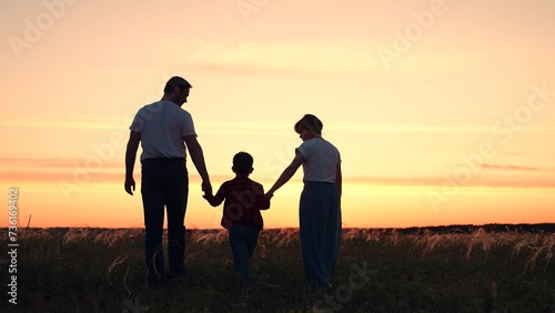 Dad mom hold their child hands in grass field. Parents child son walk in meadow. Happy family mom dad child walking in park on summer evening. Parents and son in nature. Family holidays in outdoors