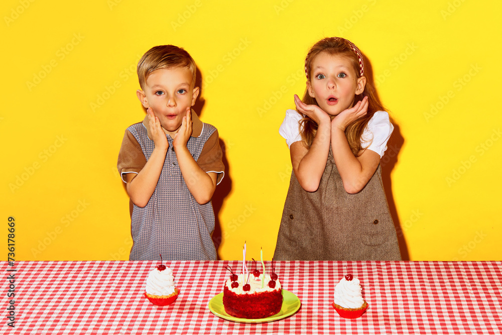 Little boy and girl, children, boy and girl standing near table with cakes with shocked face against yellow background. Excited faces. Concept of childhood, birthday celebration, family, fun, food Little boy and girl, children, boy and girl standing near table with cakes with shocked face against yellow background. Excited faces. Concept of childhood, birthday celebration, family, fun, food