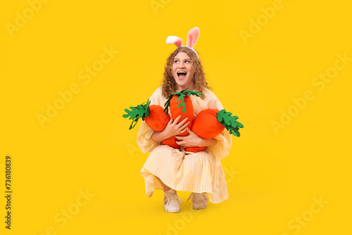 Beautiful young shocked woman in bunny ears with carrot-shaped toys on yellow background. Easter celebration